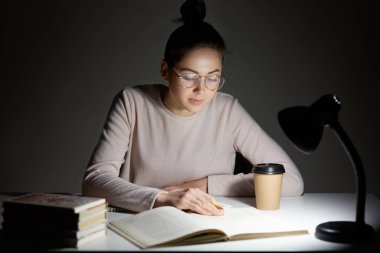 Busy female teenager reads book, uses table lamp, prepares for final examination, sits at desktop, wears optical round glasses, poses against dark background. Reading in evening. Studying concept clipart