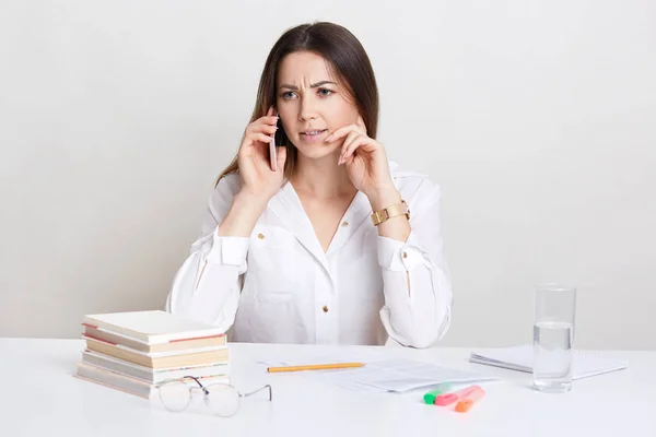 Displeased female professor talks on cell phone, wears white shirt, discusses something with indignant expression, has spectacles, pile of book, glass of water on desk. People, business, career