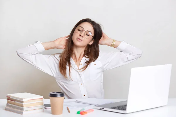 Tired female stretches at work place, wears white shirt and spectacles, feels overworked, sits in front of opened laptop, drinks takeaway coffee to feel refreshed, isolated over white background