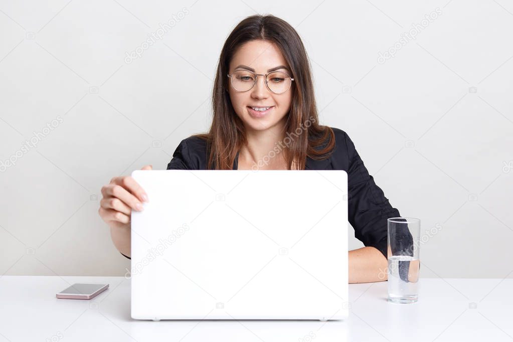 Satisfied young female sits in front of opened laptop computer, watches webinar, thinks about creating new web design. wears round spectacles for good vision, drinks water, sits indoor alone.