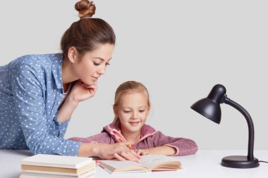 Horizontal shot of serious young mother teaches her small light haired daughter to read, shows something in book, pose at desktop with literature and readinglamp, isolated over white background clipart