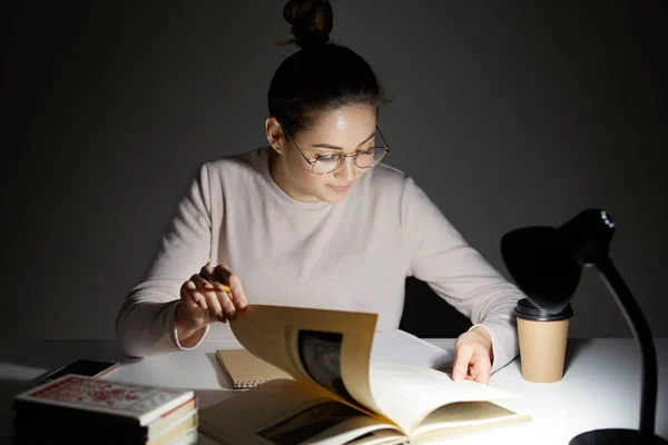 Photo of serious female sudent turns over pages in book, searches necessary information for writing course paper, wears round big spectacles, sits at table with notebook and textbooks, drinks coffee