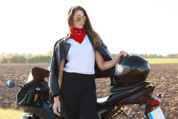 ファッショナブルな女性バイカーの水平ショット レザー ジャケット サングラス 赤いバンダナを身に着けている Motorrbike に近いポーズ 安全な乗馬のヘルメットを使用して モデルの屋外ストリート ライフ — ストック写真