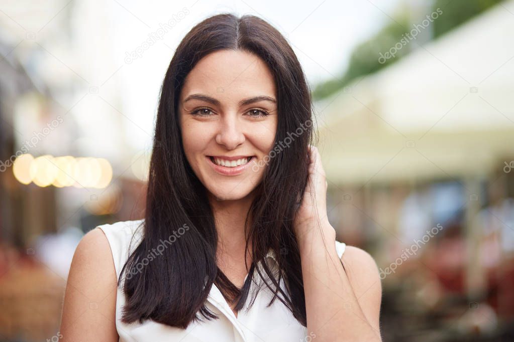 Headshot of pleased brunette female looks happily at camera, poses outdoor, enjoys spare time, models in open air against blurred background, demonstrates her natural beauty. Lifestyle concept