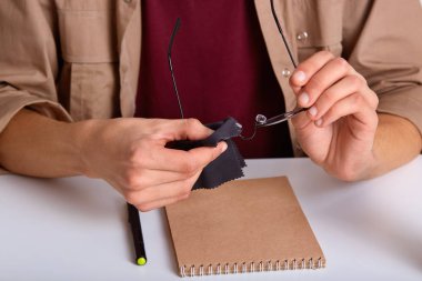 Kırpılan shot tanınmaz erkek bej gömlek, siyah mendil ile yuvarlak gözlük mendil, defter ve kalem ile beyaz masa başında oturur. Odak gözlük ile el mans. Çalışma kavramı