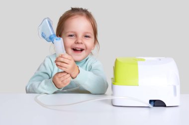 Photo of small child laughs happily, does inhalation, cures asthma, dressed in light blue turle neck sweater, isolated over white background. Therapy with inhaler. Kid with respiratory problem clipart