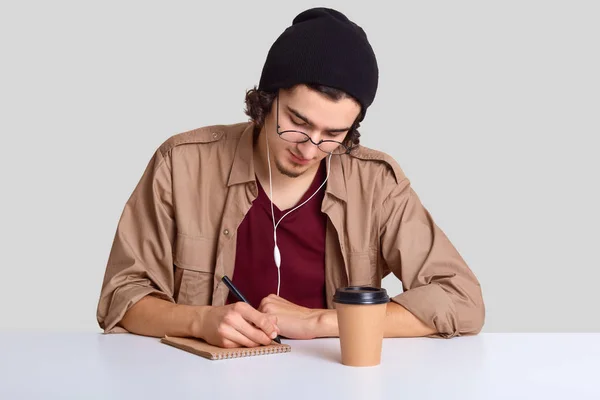 Fashionable male teenager listens music with earphones, has inspiration for writing essay, makes records in notepad, drinks coffee, isolated over white background, listens training seminar online.