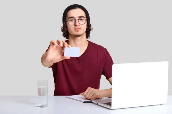 自己保証男性フリーランサー開いたノート パソコンの前に座っている ノートを作る あなたの広告のための空白のカードに手を伸ばす 電子デバイスと分離水のガラスでデスクトップでポーズ — ストック写真