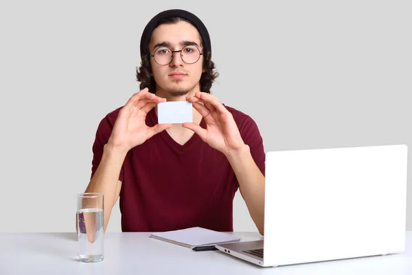 深刻な Startupper の屋内撮影丸いメガネを身に着けている 自信の表情には 空白の白いカードを保持している 水を飲む ラップトップ コンピューターを使用して 感じるのどが渇いて水を飲む オンラインの作品 — ストック写真