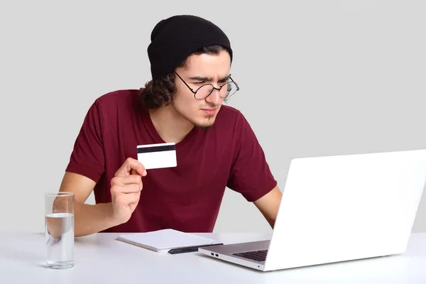 严肃的男人戴着黑帽 圆圆的眼镜 在便携式笔记本电脑上仔细地看着屏幕 做银行数据的验证 拿着塑料卡 隔着白色的背景 银行理念 — 图库照片