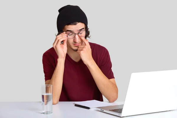 疲れた男 Rubbs 身に着けているメガネの写真目のラップトップ コンピューターで長い仕事の後痛みがある カジュアルな シャツを着ている コース作業の準備 白い壁で分離された現代のポータブル ラップトップを使用して — ストック写真