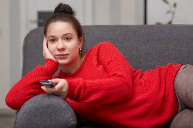 Hamile kadın evde Tv izliyor, uzaktan kumanda tutar, film sahiptir, kanepeye, yalan olan tembel gün, monitöre odaklı. Gebelik, dinlenme ve yedek zaman kavramı. Gelecekte anne çocuk bekliyor