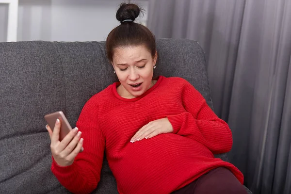 Omg, childbirth starts! Puzzled pregnant woman keeps hand on belly, has birth pain, wears red sweater, holds modern cell phone, calls ambulance, poses at cozy sofa. Future mom feels pain inside