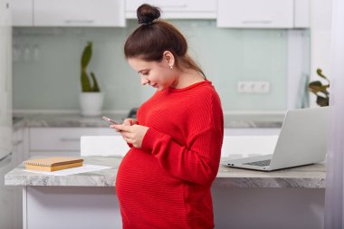 Yana doğru shot esmer genç kadının kocası hücresel, alır bildirim üzerinde metin ileti türleri, mutfak yalnız teşkil etmektedir, taşınabilir dizüstü bilgisayar ve kitap ile beyaz masa yanında duruyor.