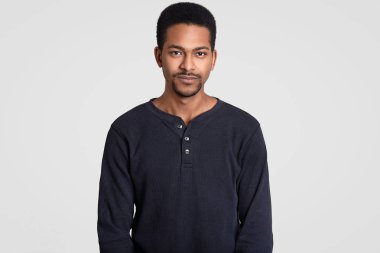 Bel küçük sakal, kısa kıvırcık saçlı zenci adam çekim kadar ciddiye kamera, beyaz duvara izole gündelik kıyafeti giymiş düşünceli yüz ifadesi ile bir şey hakkında düşünüyor görünüyor.