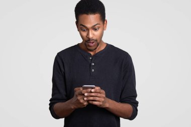 Şaşırmış siyah adam tutar modern cep, alır beklenmeyen metin iletisi, surfes web sayfası, rahat kıyafetler giymiş beyaz arka plan üzerinde izole şok, bağlı kablosuz intenet hissediyor
