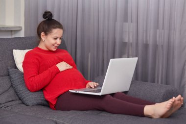 Yerli atmosphhere, dizüstü bilgisayarda tuş takımı bilgi memnun koyu saçlı hamile genç kadının kapalı atış sahiptir, oturur yüksek kablosuz bağlı kanepeye, yalınayak hızlı internet