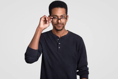 Waist up shot of handsome self confident young male manager, keeps hand on rim of spectacles, wears black casual jumper, tries to make decision or solve problem, isolated over white background clipart