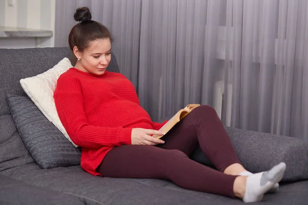 満足の暗い髪の女性 子供を出産する予定だ レギンス 赤いジャンパーを着て 本を保持している Inreresting ストーリーを読み取り クッションに傾いて リビング ルームのソファーでポーズします — ストック写真