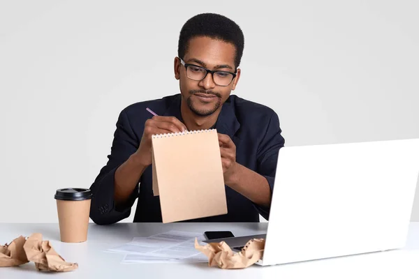 スパイラル メモ帳でアイデアを書き込み 作成新しい記事に深刻な黒人男性ジャーナリスト作品のショット ペーパー コーヒーに囲まれたラップトップ コンピューターで動作 テーブルの上の創造的なカオス — ストック写真