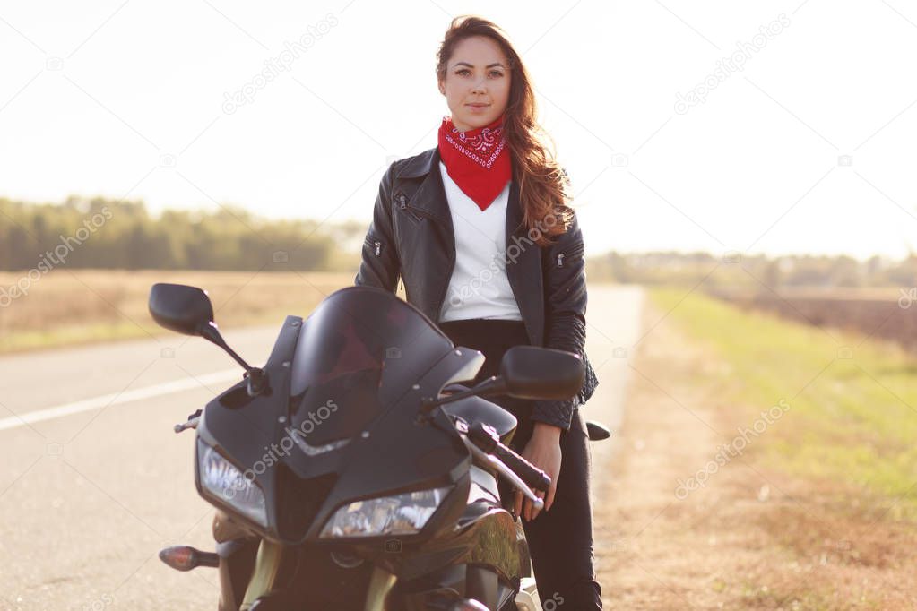 Female motocross racer dressed in black leather jacket, poses on her motorcycle, has adventure in countryside, likes risky sport, looks directly at camera. People, motorcycling and extreme concept