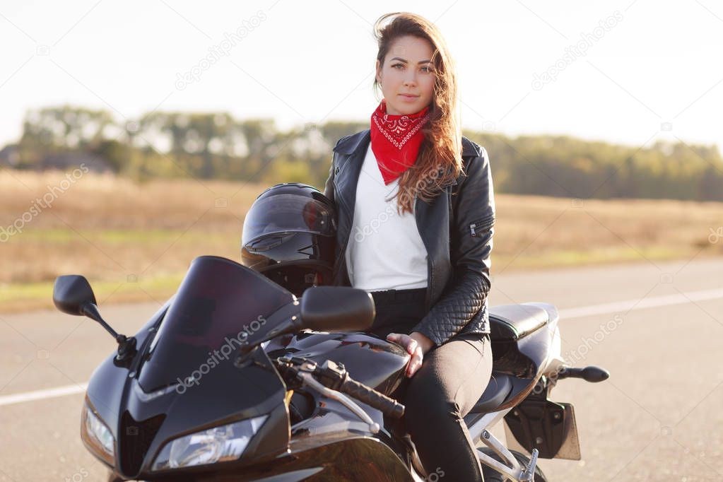 Outdoor shot of pretty woman biker wears red banadana and leahter jacket, holds helmet, sits at fast black motorbike, poses over road background, has red stylish bandana on neck. Motorcycling