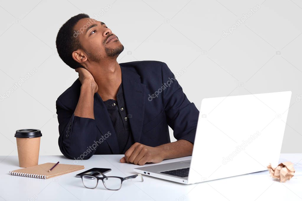 Dreamy dark skinned male worker has pain in neck, keeps gaze upwards, works for long time at desktop, prepares new publication on business theme, drinks hot beverage, isolated over white wall