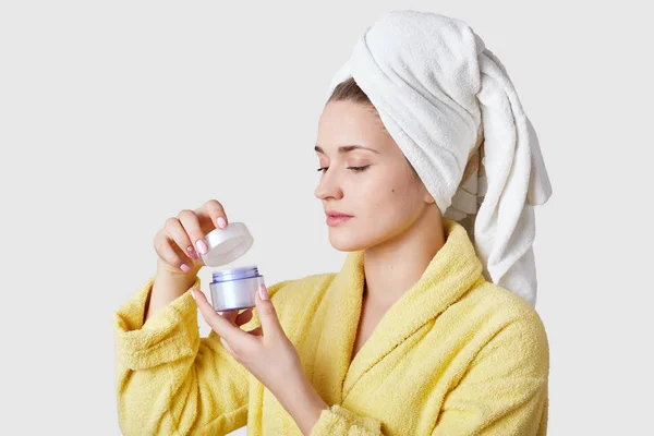 Sideways shot of healthy young European woman wears white towel and yellow bathrobe, holds cream, going to have beauty treatments, isolated over white background. Wellness and skin care concept