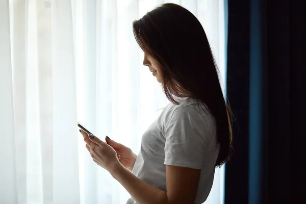 シャツで暗い髪の若い女性の横向きのショット メールをチェック 自宅の自由な時間を楽しんで 窓の近くに立つ ワイヤレス インター ネットに接続されている現代の携帯電話を使用して テキスト メッセージを送信 — ストック写真