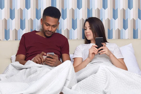 图为上瘾的混血可爱的家庭夫妇手里拿着现代手机 冲浪社交网络 坐在舒适的床上 床上用品下 连接无线网络 在家休息 — 图库照片