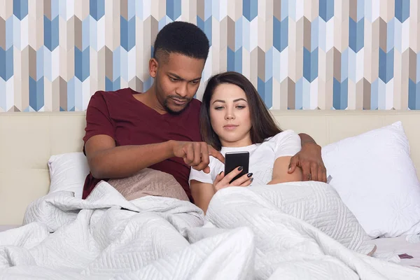 多文化の若い女性と男性は インターネットのウェブストアで何かを選択 携帯電話の画面に黒い肌の男性のポイントは 妻にこの商品を購入して 居心地の良い寝室のベッドで一緒にポーズを示唆しています — ストック写真