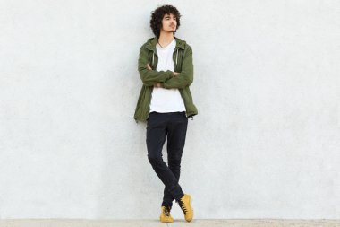 Moda giysiler giymiş düşünceli kıvırcık adam vurdu, tutar silah katlanmış, bakışları, beyaz stüdyo duvardan izole bir şey hakkında düşünüyor tutar. İnsanlar, stil, gençlik ve güven kavramı