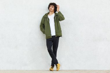 Moda giysiler, pozlar beyaz stüdyo duvar karşı düşünceli ifade, çizikler kafa, kıvırcık çekici adamla fotoğrafını giymiş yalnız düşünüyor hakkında bir şeyler hissediyor. İnsanlar ve stil kavramı