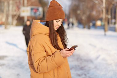 Şık şapka ve gevşek ceket giymiş kadın genç yana shot cep telefonu, surfes sosyal ağlar tutar ve video izliyor, karlı şehir arka plan üzerinde teşkil etmektedir. Online iletişim