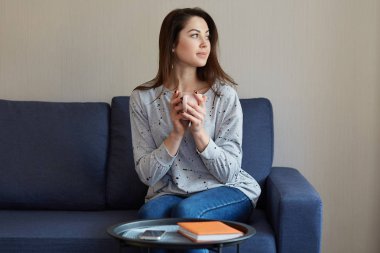 İnsanlar, ev concept. Düşünceli oldukça Avrupa kadın sıcak içecek, kazak ve kot pantolon giymiş bir tutar, odaklanmış bir yana, kanepe cep telefonu ve yakın kırmızı Not defteri ile yuvarlak masa oturur