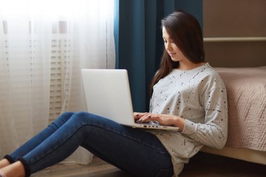 Çekici kadın fotoğrafı şık kazak ve kot pantolon giymiş, dizüstü bilgisayar diz tutar, monitöre odaklı bilgi, klavye, kablosuz internet'e bağlı yatak, yakın katta oturur