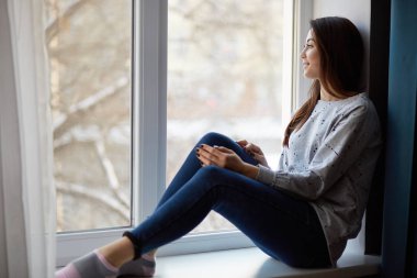 Düşünceli rahat kadın modeli kapalı atış rahat jumper, kot pantolon giymiş pencere evde oturur ve çorap, kış aylarında güzel görünümü penceresinden hayran, hafta sonu veya gün vardır