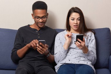 Sürpriz güzel genç kadın yakınındaki modern cep telefonu, derili haber online, karanlık adam giyer gözlükler, şok edici okuma oturur tutar, kablosuz internet bağlantısı evde zevk, rahat kanepe otur