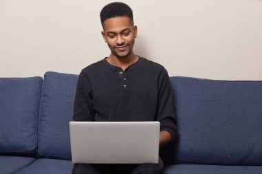 Memnun koyu tenli adam fotoğrafı sakal vardır, siyah kazak giyer, rahat kanepe pozlar, kablosuz internet, okuma bilgi proje çalışması için bağlı uygulamayı yükler.