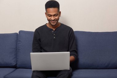 Memnun Afro-Amerikan bağımsız açılan dizüstü bilgisayar önünde kanepede oturuyor, uzak iş yok, gündelik kıyafeti giyer, Aile içi atmosfer, yüksek hızlı internet bağlantısı sahiptir. Teknoloji