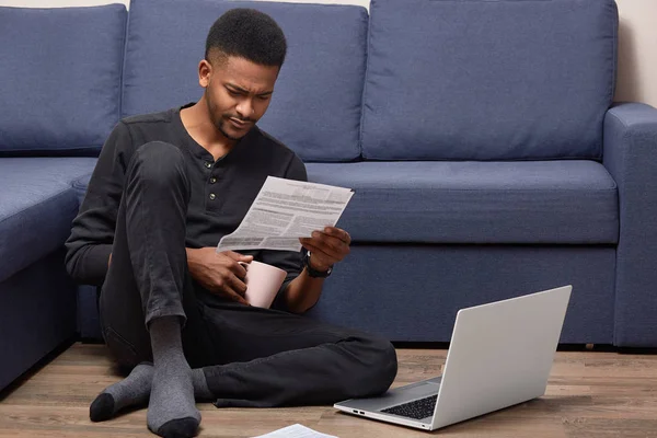 Young prosperous black entrepreneur has attentive look at document, studies contract terms, wears casual clothes, works with laptop computer, poses at floor near sofa in living room, works on distance