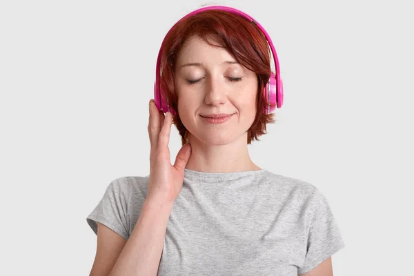 満足している若い女性のヘッド ショット ヘッドフォンを身に着けている お気に入りの音楽に耳を傾け 喜びから目を閉じて 大きな音を楽しむカジュアルな シャツ 白い背景で隔離に身を包んだ 趣味の概念 — ストック写真