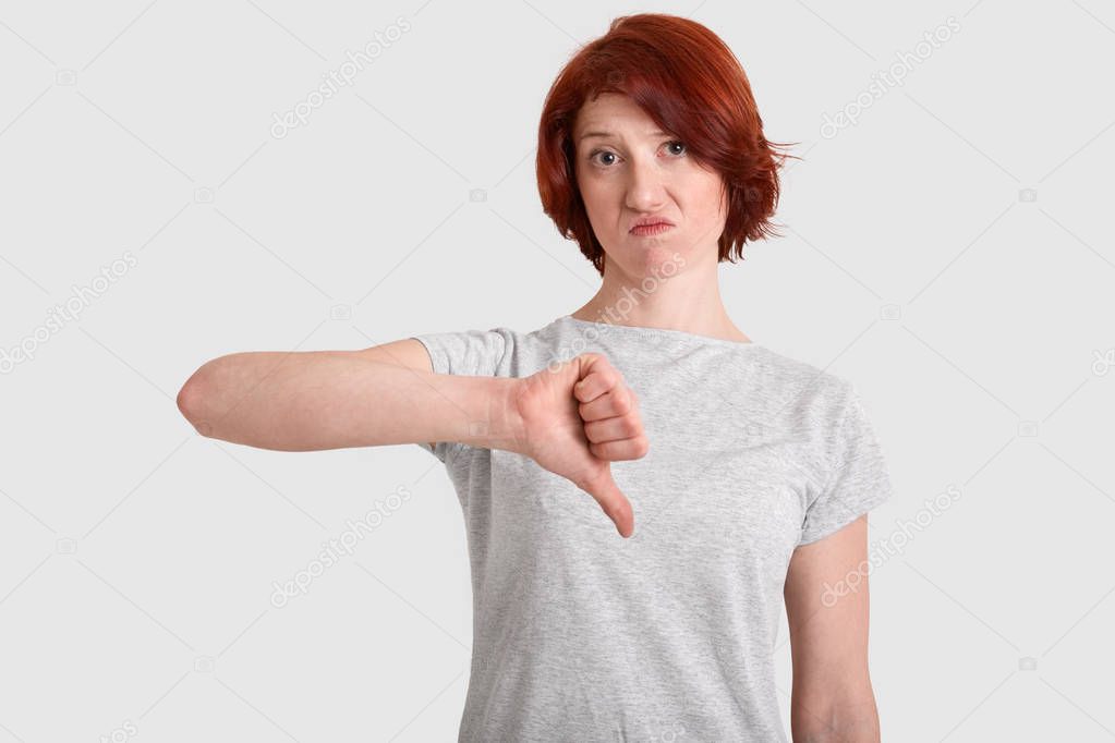 Displeased red haired woman keeps thumb down, frowns face, doesnt like something, dressed in casual t shirt, models over white background. Photo of young ginger lady shows dissatisfaction gesture.