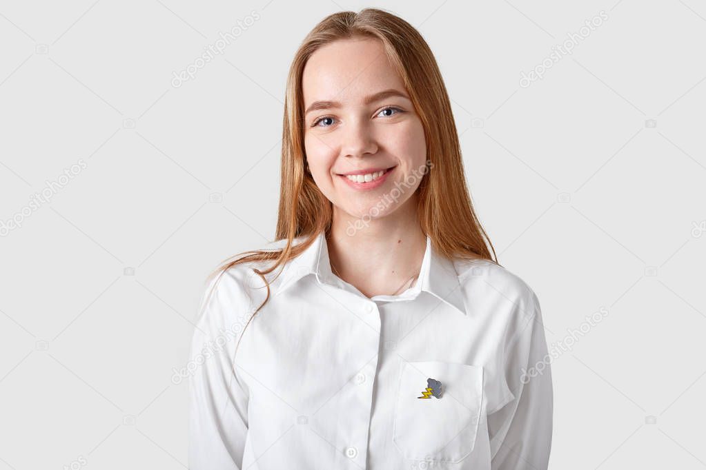 Smiling charming young girl with blue eyes, toothy smile, dressed in white shirt, has long hair, isolated over white background. Pleased European lady expresses positive feelings good emotions