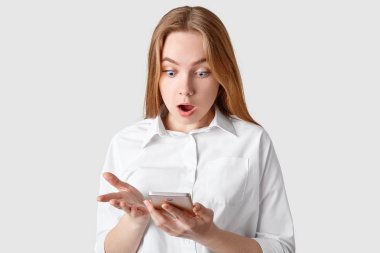 Şok korkunç kadın akıllı telefon ekranında Haberler, beyaz arka plan üzerinde izole şık gömlek giymiş şok edici okuma dik dik bakıyor. Kapalı atış ile modern becerikli alet şaşkın kız.