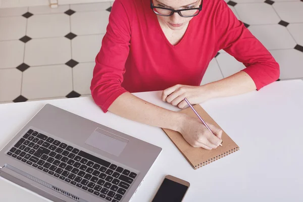 Cropped top view of busy female writer makes notes in spiral notepad, wears transparent glasses and red sweater, focused in screen of laptop, sits at white desk, rewrites information from internet