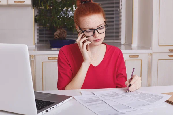 Serious busy freckled woman with ginger hair, wears spectacles, underlines information in papers, has phone conversation, sits in front of opened laptop against kitchen interior. Paperwork concept