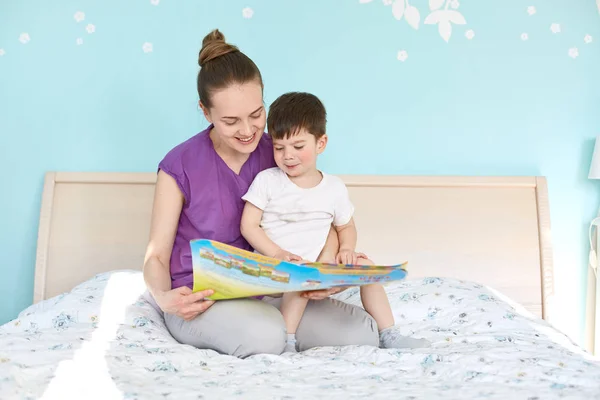 Indoor shot of affectionate woman and small boy read interesting story from book, sit at bed, enjoy calm atmosphere, smile positively. Mother and little male child pose in bedroom. Education