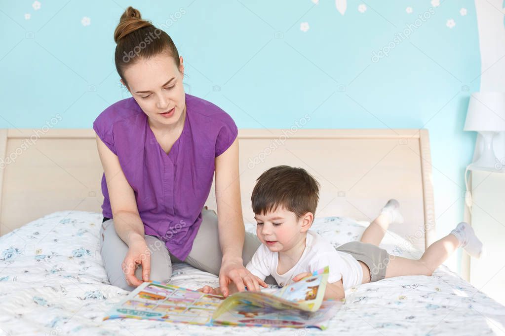 Caring young mother reads magazine with pictures for children to her small son, pose together at bed against cozy interior and blue wall, enjoy domestic atmosphere. Mum and kid. Generation concept