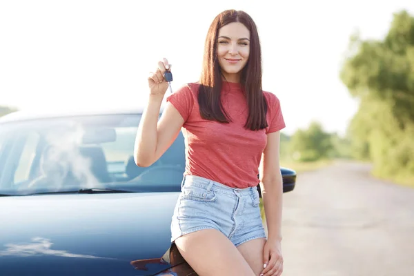 Pretty brunette young European woman shows car key, rejoices new purchase, poses near new automobile, dressed in casual t shirt and denim shorts, poses outside. Female seller advertises her vehicle